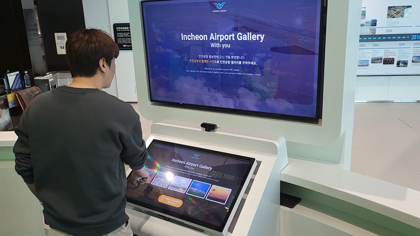 인천공항 제2여객터미널 역사관 AR 포토존은 32인치 터치 키오스크에서 배경을 선택하고 촬영하면, 55인치 전면 모니터에 그 결과가 보이고, 55인치 후면 모니터에는 촬영된 사진들이 모여 갤러리 형태로 표시되는 사례입니다.